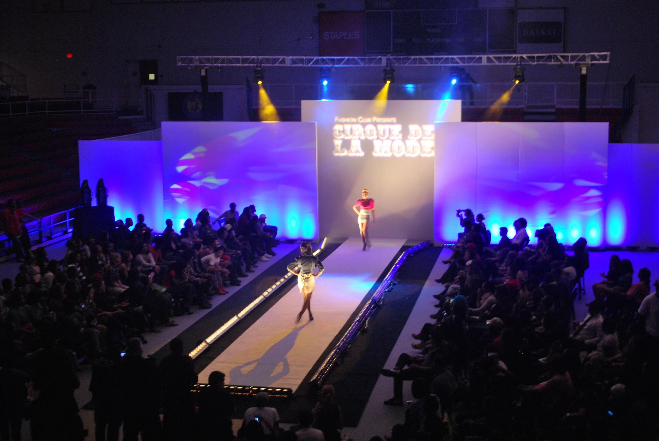 A large group of people sitting around a runway for a fashion show. Two models are currently walking the runway.