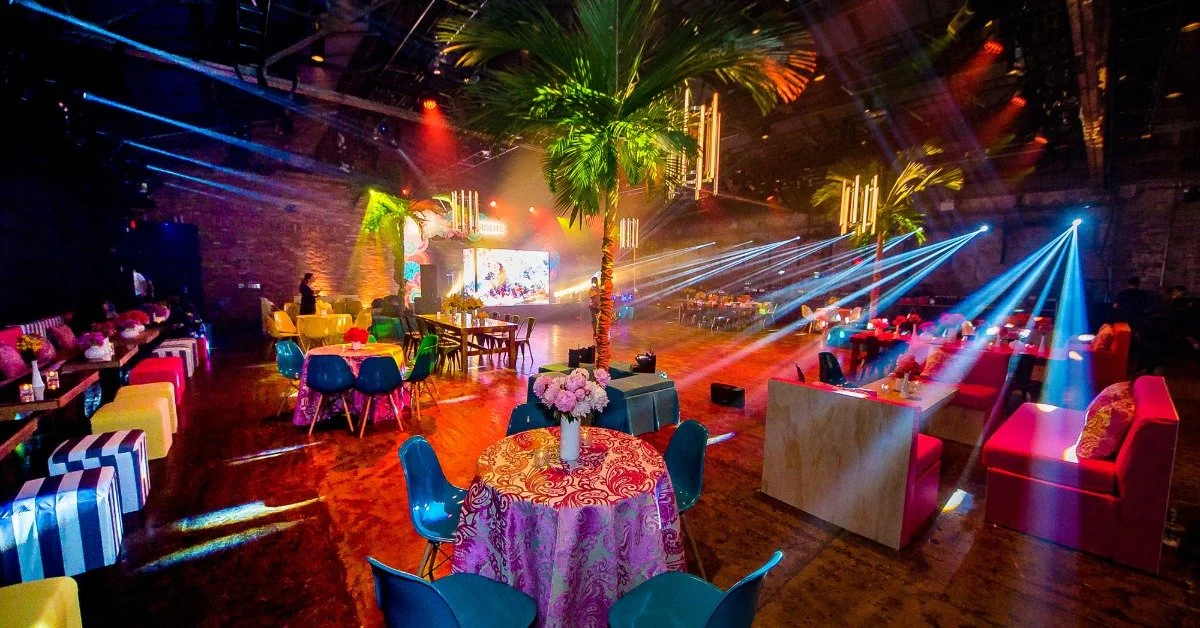 A well-decorated event space with lights streaming overhead. There are multiple tables with chairs set up around the center.
