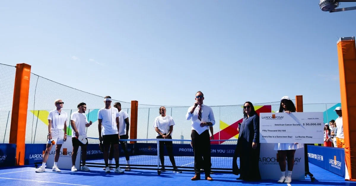 A group of people standing on a small, blue pickleball court. One person is holding a large check next to the net.