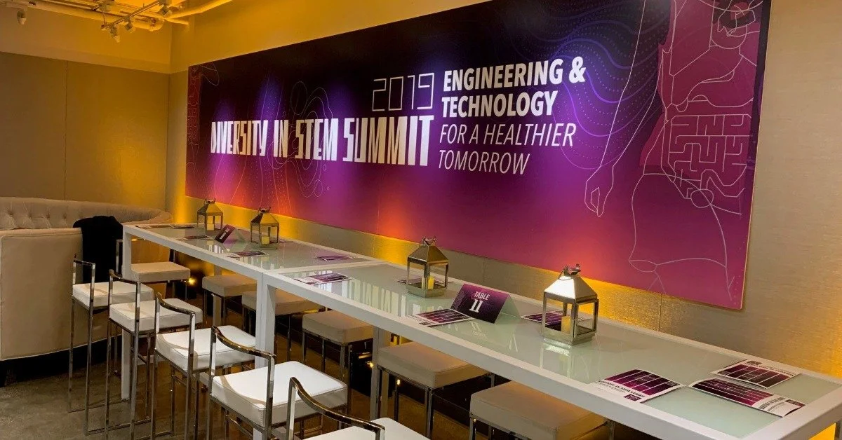 A tall table with multiple stools positioned in front of it. The wall behind it has a poster for a Diversity in STEM Summit.