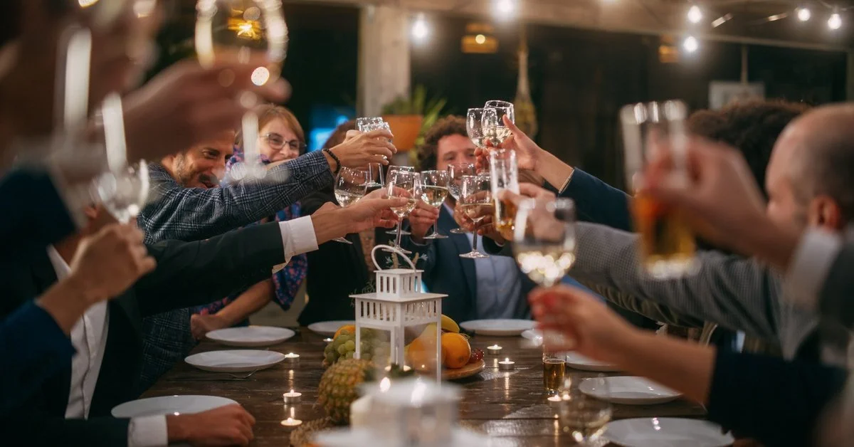 A large dinner party with many people raising wine glasses in a cheers motion. Each person has a plate in front of them.