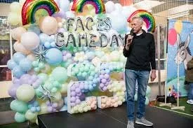 A man with a microphone standing in front of a wall with dozens of multicolored balloons attached to the surface.