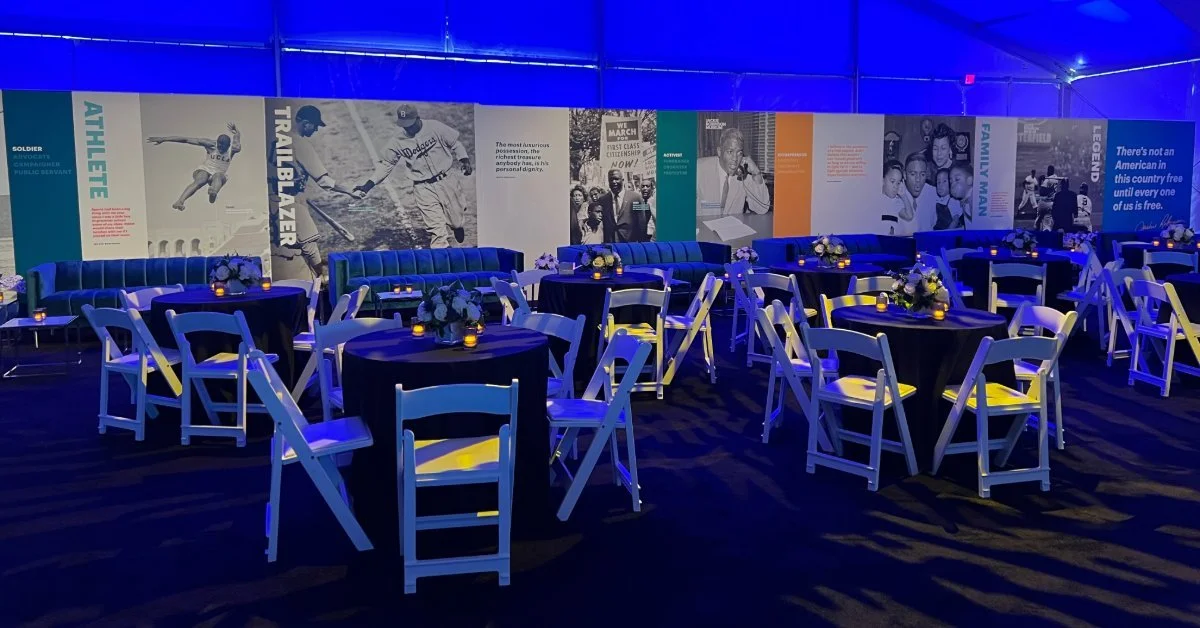 A reception space with round tables, white folding chairs, floral centerpieces, and blue ambient lighting. A large wall display featuring historical photographs and biographical text spans the background.
