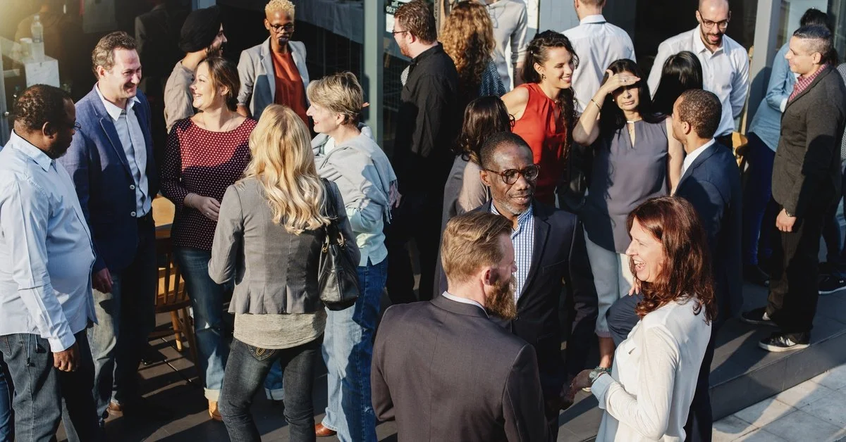 A large, diverse group of people standing together outside. Various smaller groups have formed and are talking to each other.