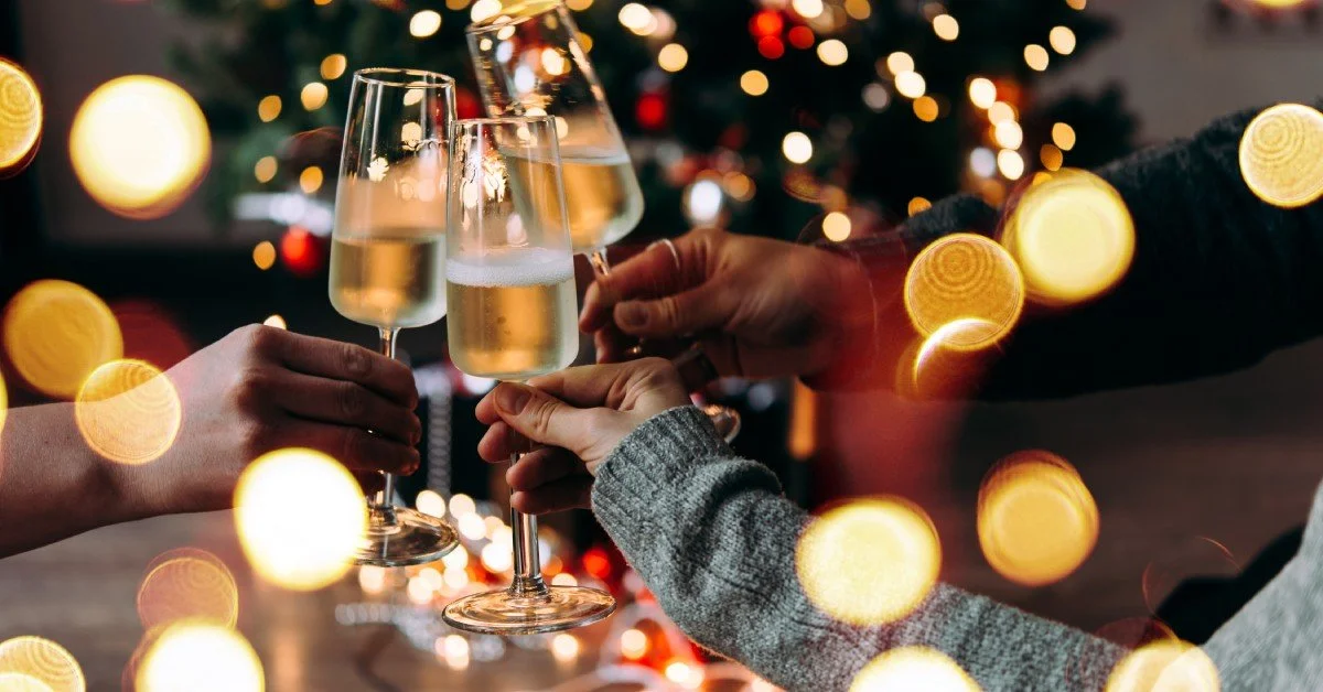 Three hands come together in a cheers motion in front of a tree with lights. Each hand is holding a glass of champagne.
