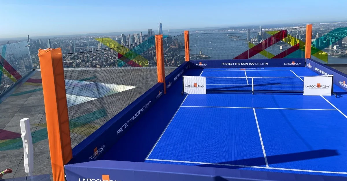 A blue rooftop pickleball court with orange posts, netting, white lines, and a city skyline in the background.