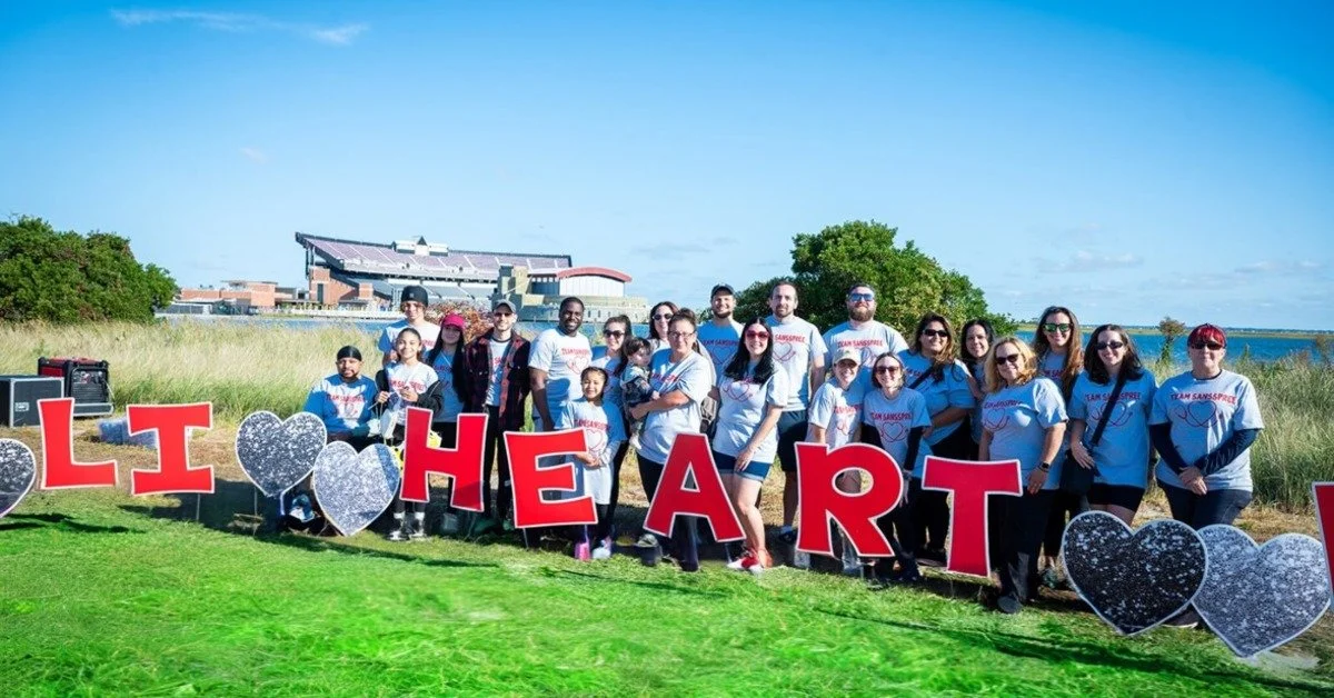 A group of people standing outdoors behind large red letters and heart shapes near grass, water, and a stadium.