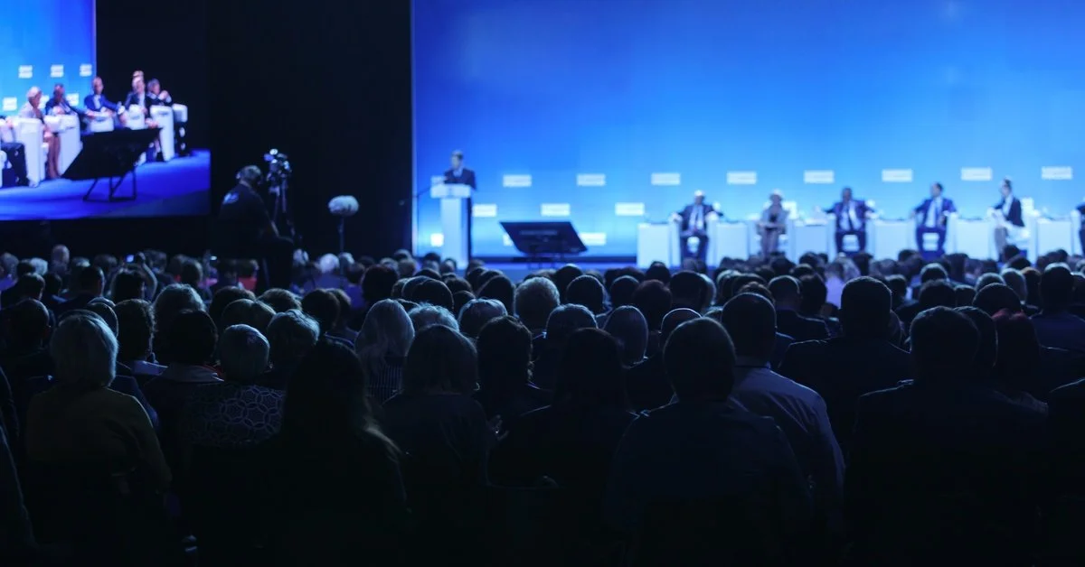 The view from the back of a crowd at a corporate event. There is a speaker at a podium near the front.