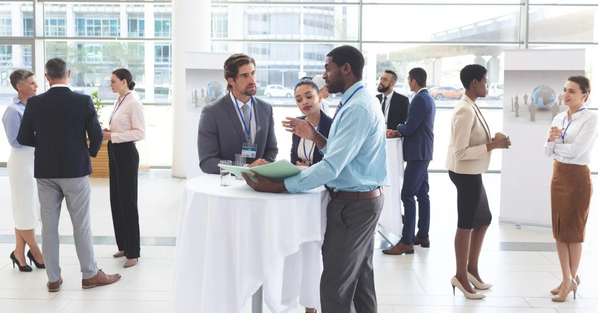 A gathering of several business people in formal dress. They are standing around tall tables with tablecloths on them.