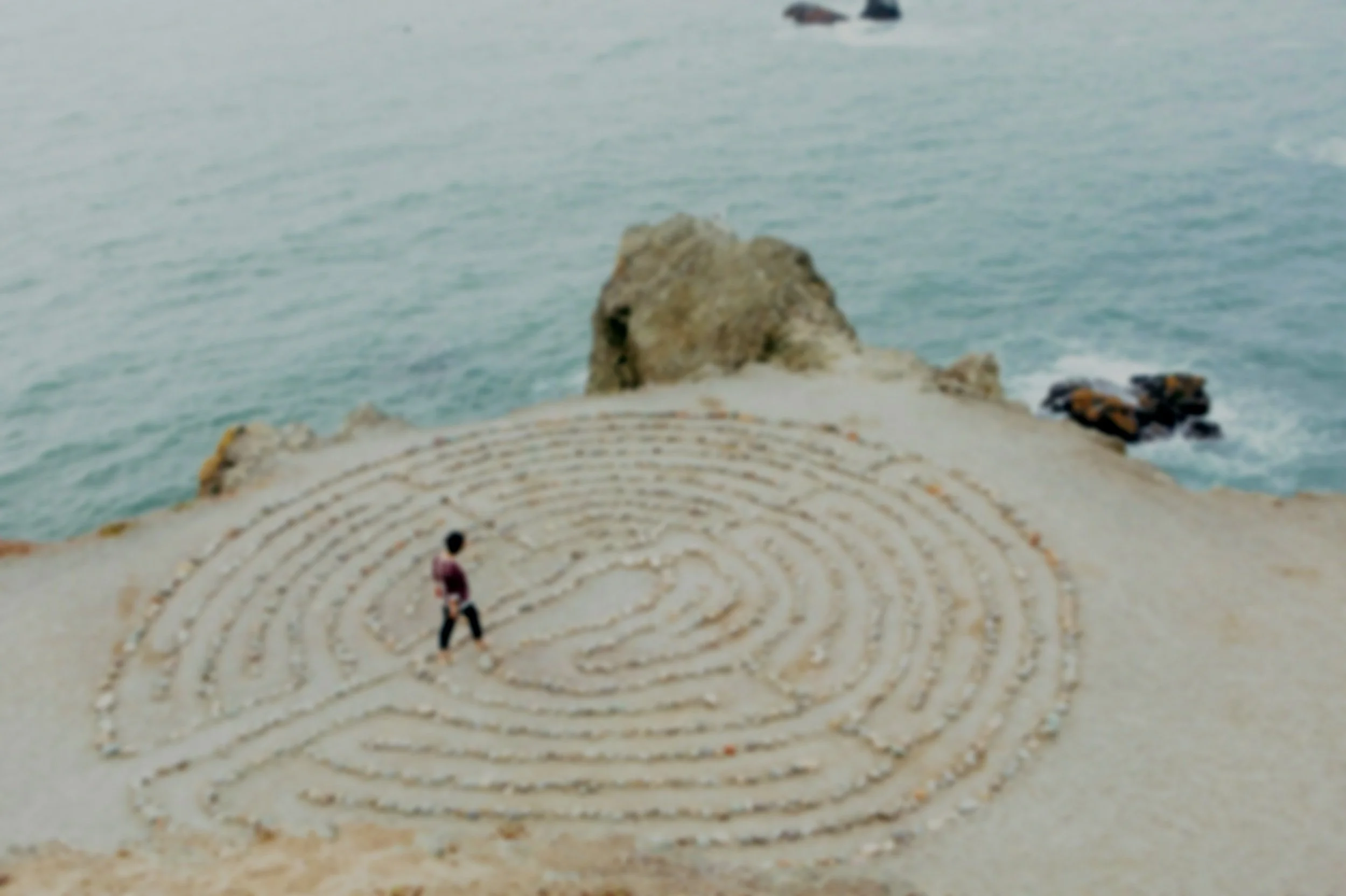 PERSON WALKING A MAZE MADE OF STONES ON A SAND BEACH WITH SEA IN THE BACKGROUND