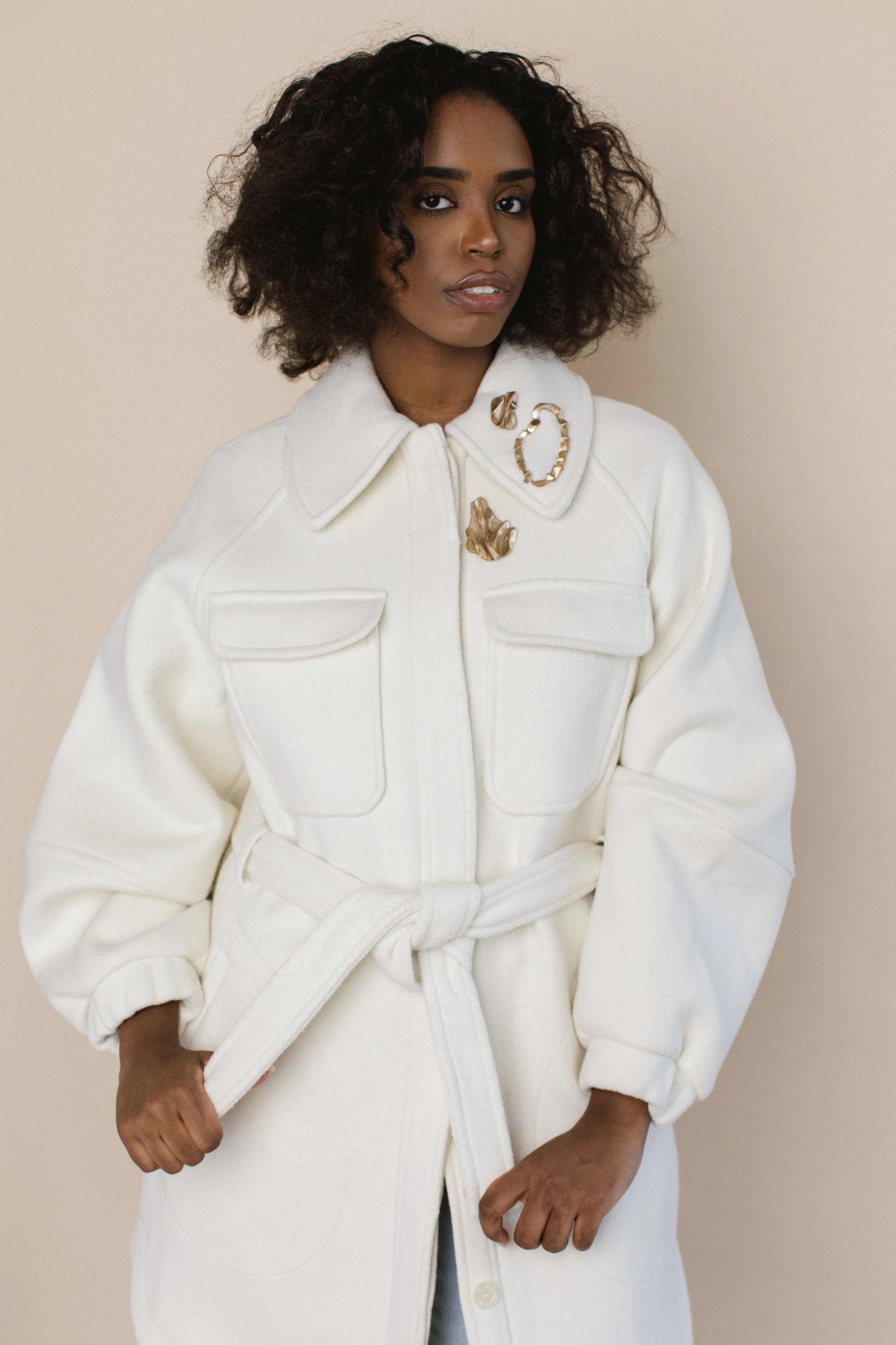 A woman wearing a cream-colored coat with large pockets and gold Be The Other brooches on the collar, standing against a neutral background.