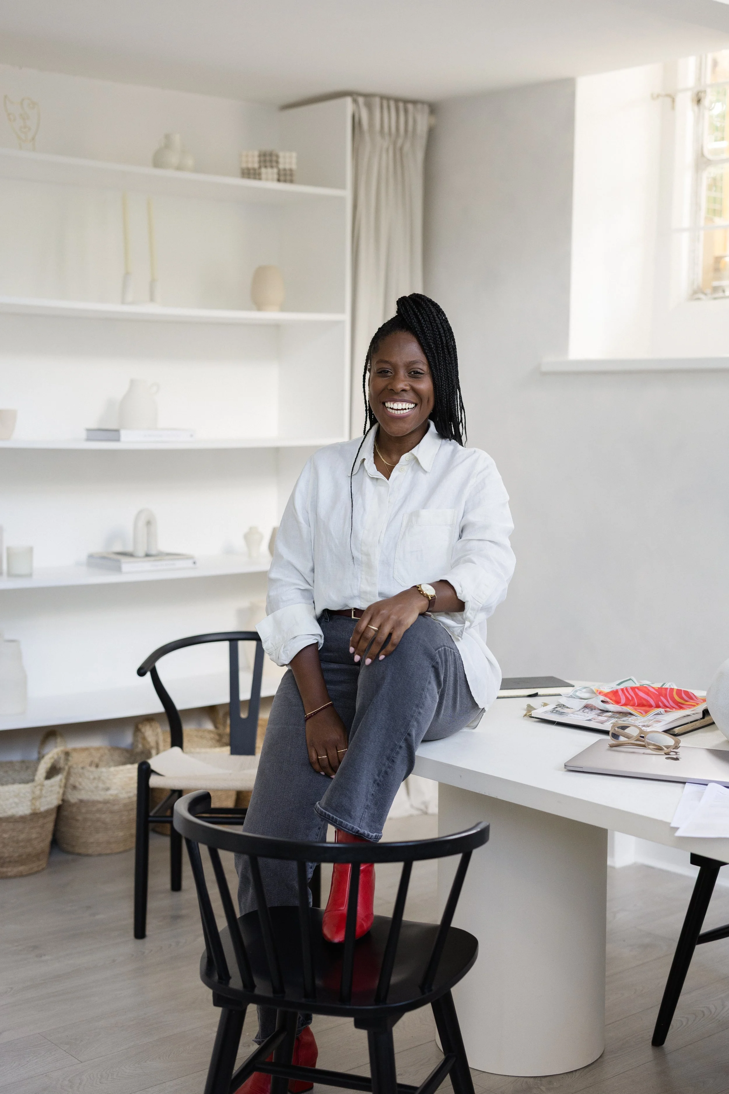 A woman with braided hair sitting on a desk, smiling, in a bright, minimalist room with white shelves and a window.