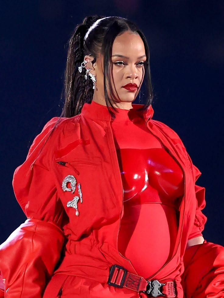 Rihanna performing on stage wearing a full red outfit with a cluster of large diamond brooch pinned to the jacket, showing her pregnancy bump and decorative accessories.
