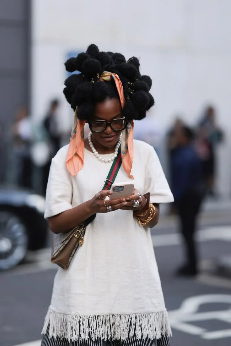 Woman wearing a white dress with tassels with a patterned silk scarf tied in her hair, with the ends flowing down the sides of her face, with oversized glasses and a vintage Gucci bag