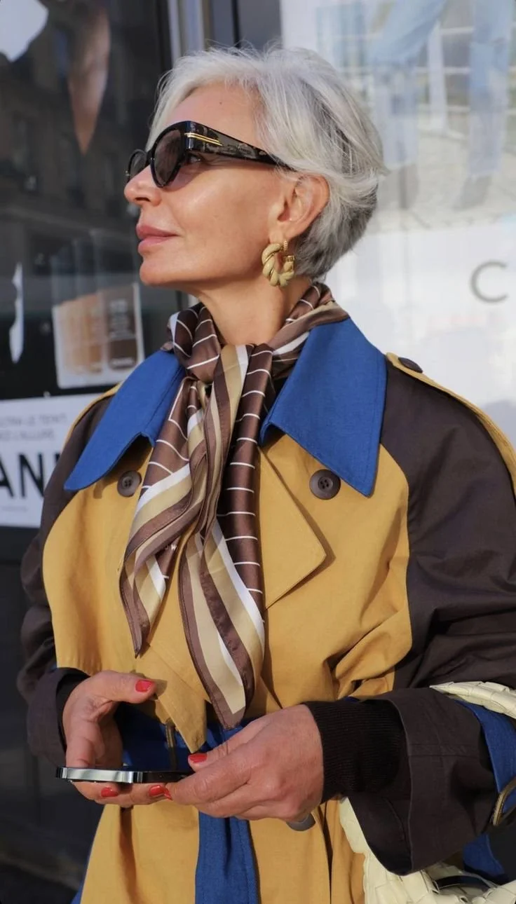Stylish woman wearing a yellow, blue, and brown trench coat with a chocolate-brown silk scarf tied loosely around her neck like a tie, with oversized sunglasses
