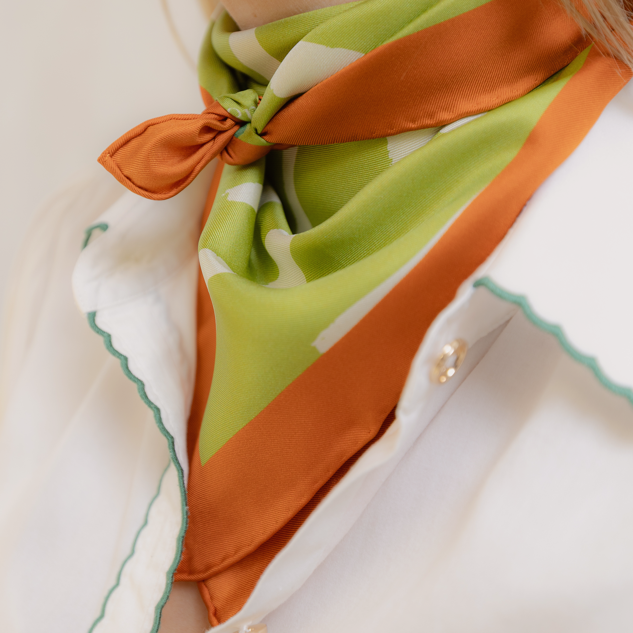 Close-up of a green and orange scarf tied in a knot around a woman's neck, featuring orange, green, and cream abstract patterns with a silky textile finish.