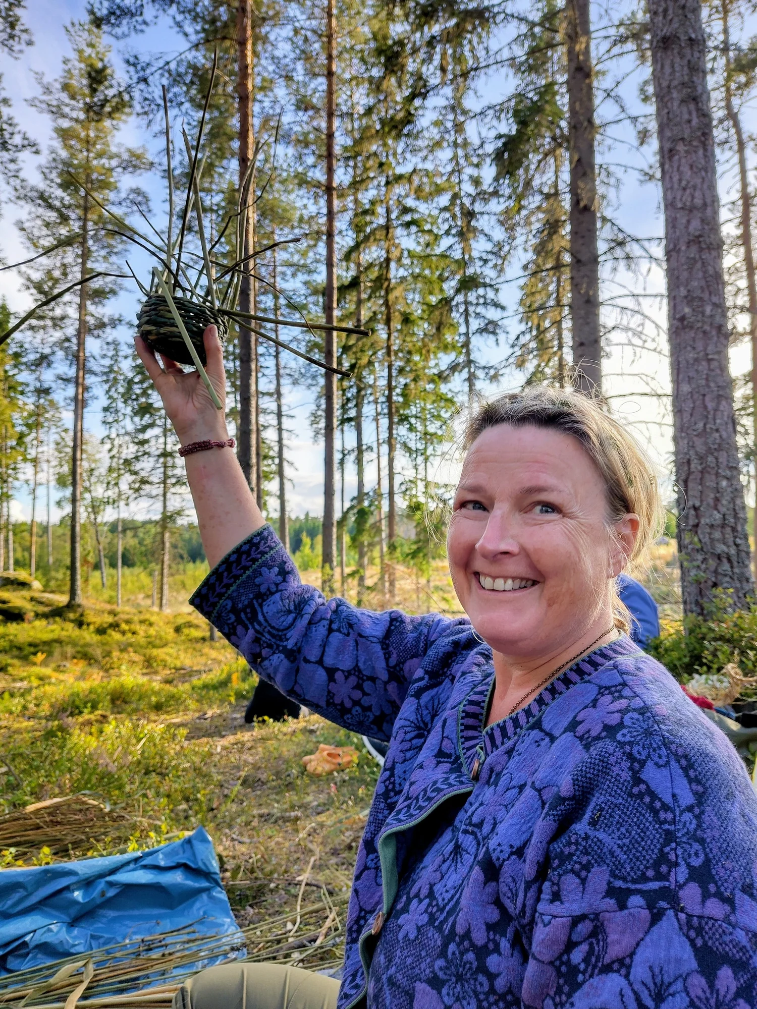 En leende deltagare i solig tallskog håller stolt upp sin påbörjade korg