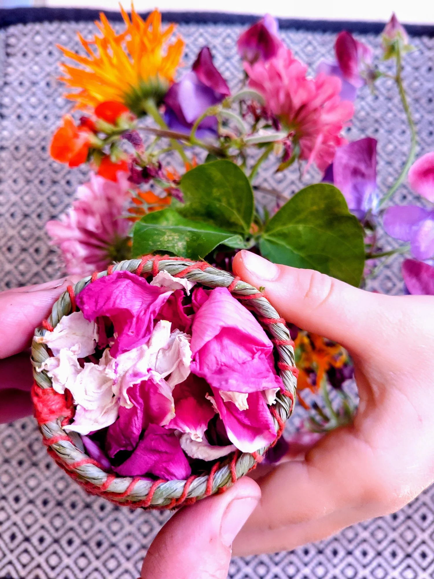 Händer håller en liten flätad korg fylld med rosenblad, med sommarblommor i bakgrunden
