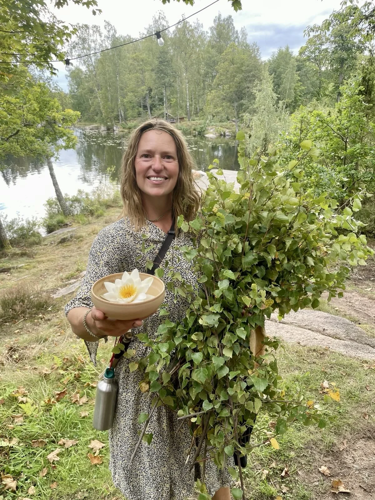 Disa ler mot kameran vid sjön, med en näckros i en keramikskål i ena handen och en famn björkris i den andra