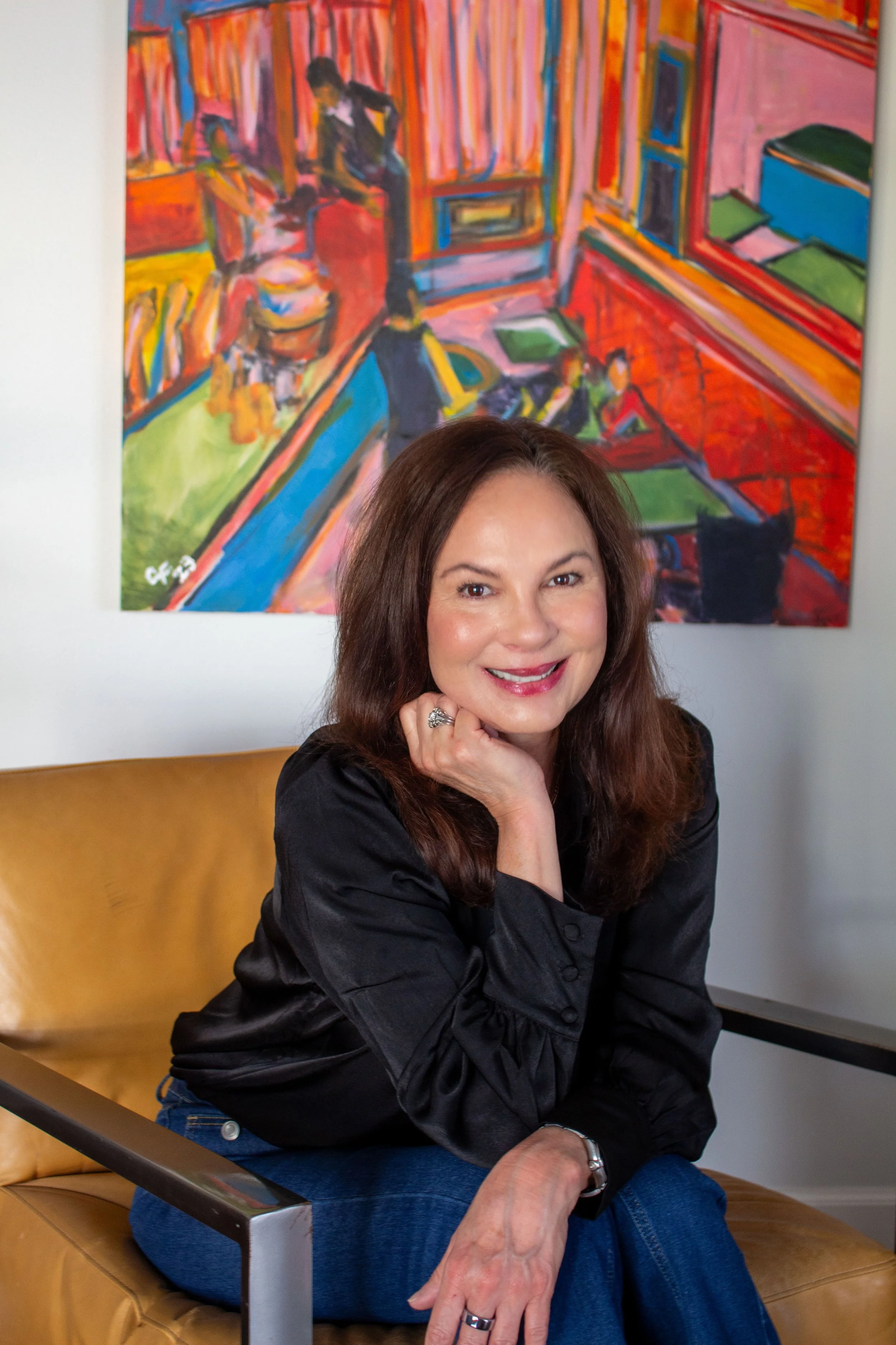 A woman with long brown hair and a black blouse smiling while sitting on a tan leather chair in front of a colorful abstract painting.