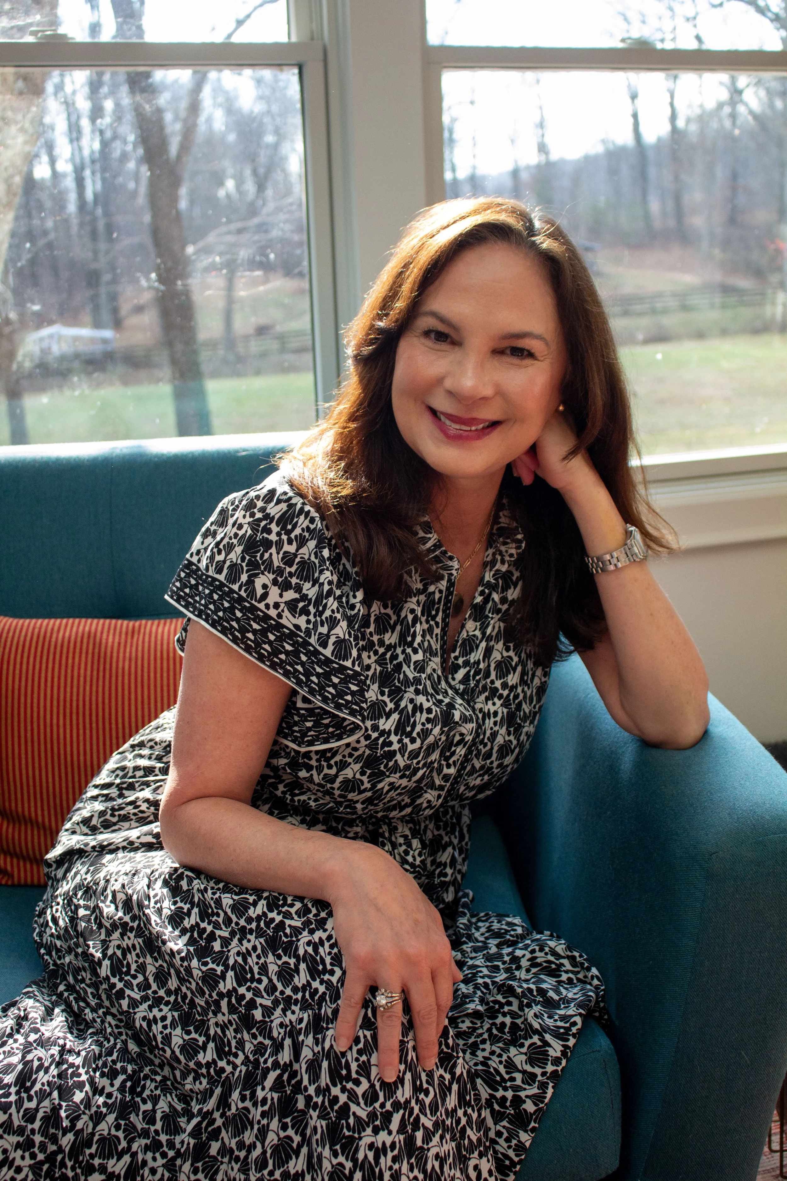 A woman with brown hair sitting on a teal couch near a window, smiling and resting her head on her hand, wearing a black and white patterned dress.