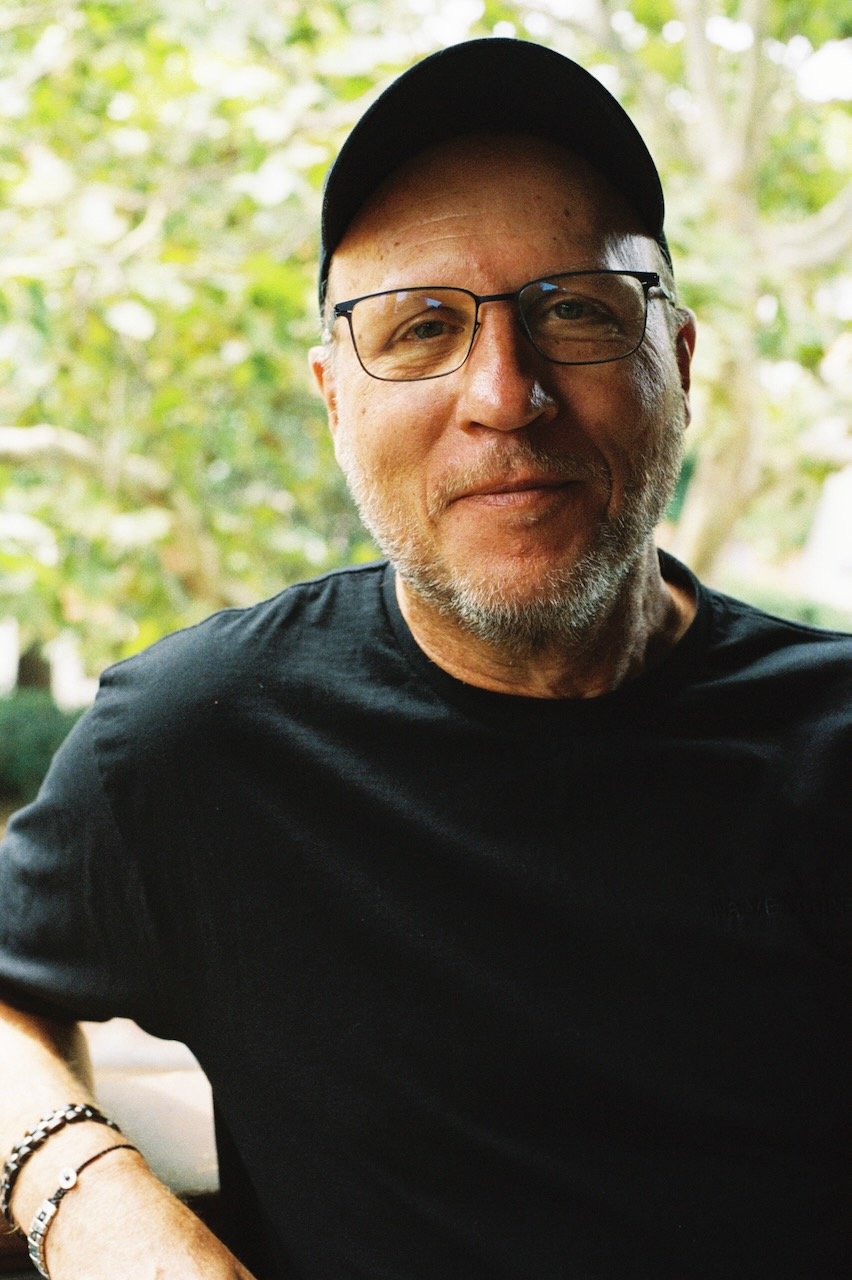 Alan leaning with elbows on wall behind him, in black ball cap, black t-shirt, trees in background.