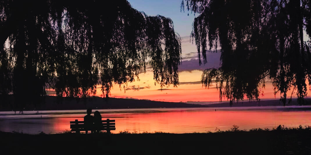 Silhouettes of two people sitting on a bench by the water during a sunset with colorful sky and overhanging tree branches.