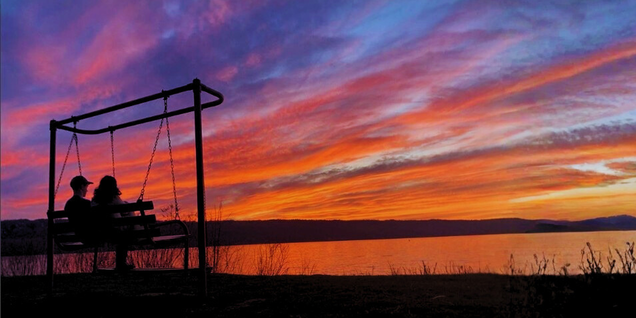 Silhouettes of two children sitting on a swing bench near a lake during a colorful sunset.