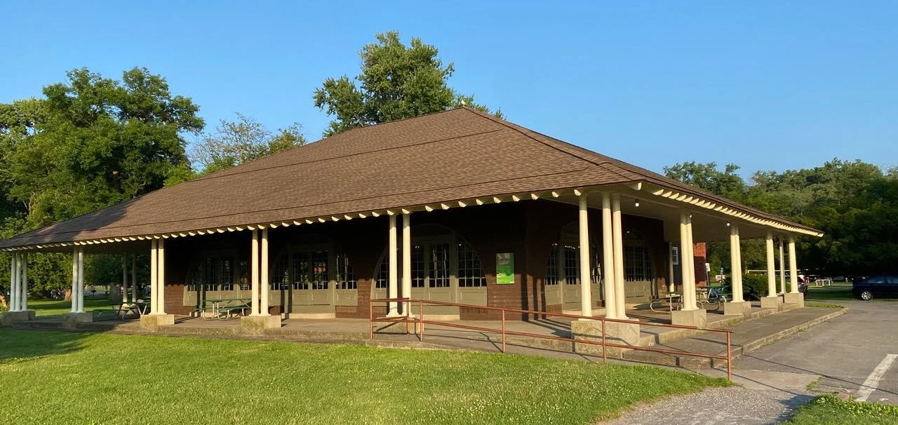 A single-story brick building with a brown shingled hip roof, covered porch with white columns, and large windows, surrounded by green grass and trees.