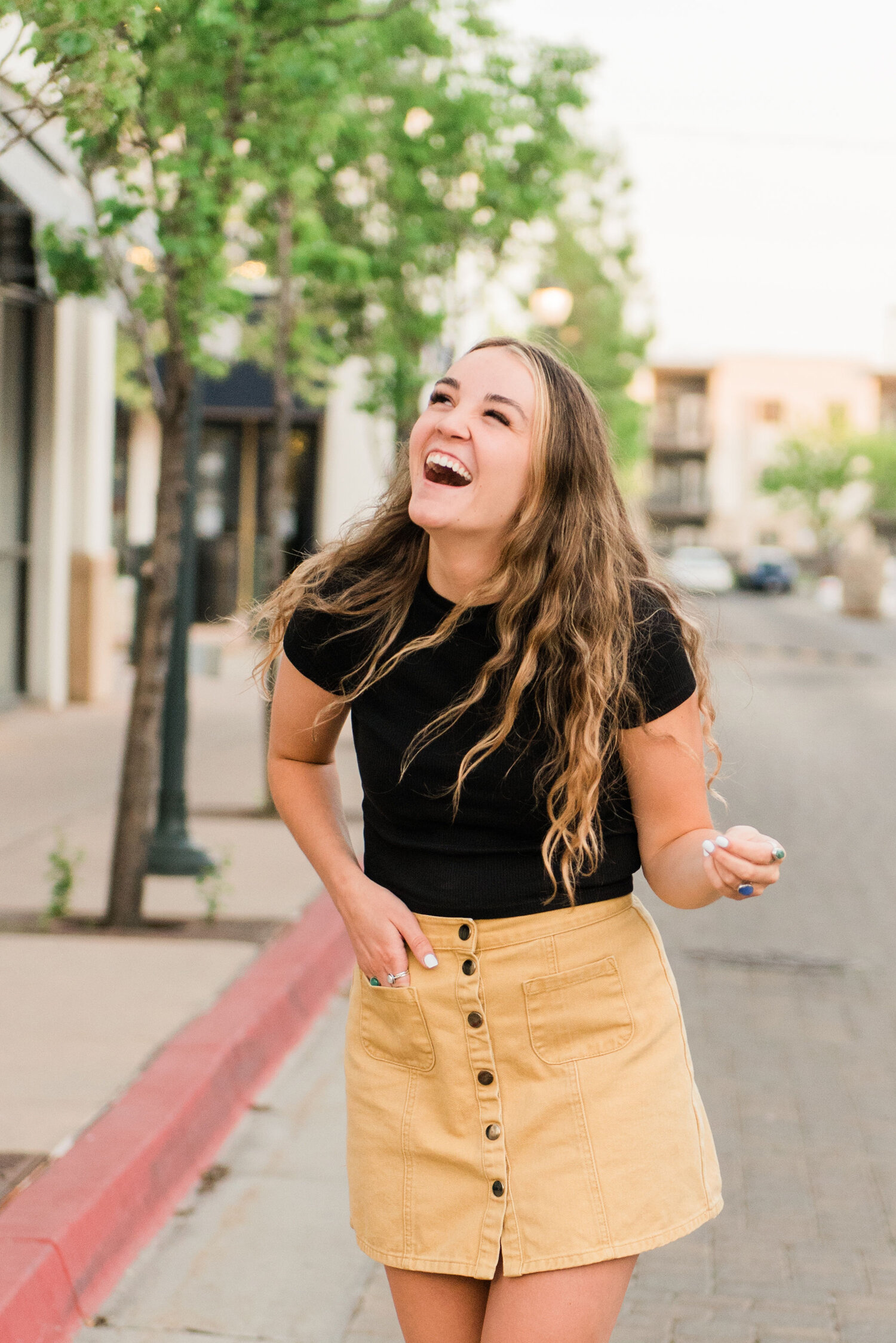  Jacquie Erickson Photography captures this graduating senior walking and laughing down an urban sidewalk for her senior photoshoot in Baltimore, Maryland. womens mini corduroy skirt with front buttons, womens long dark blonde crimped hair, laughing 