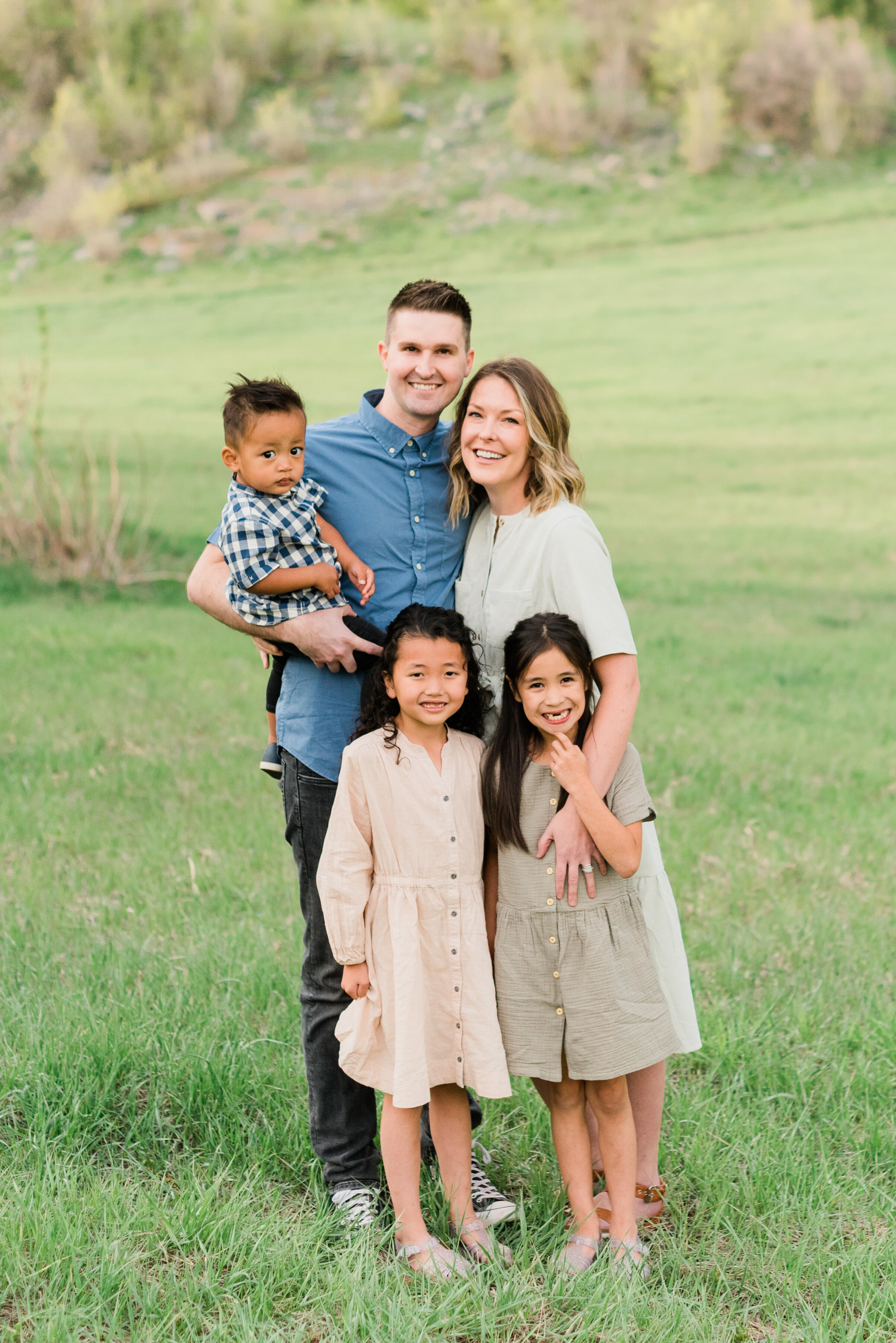  A young family of five cuddles together for a family photo in a grassy open field. family of five daughters and son multinational family  mountainside photos #jacquieericksonphotography #youngfamily #outdoorphotoshoot   