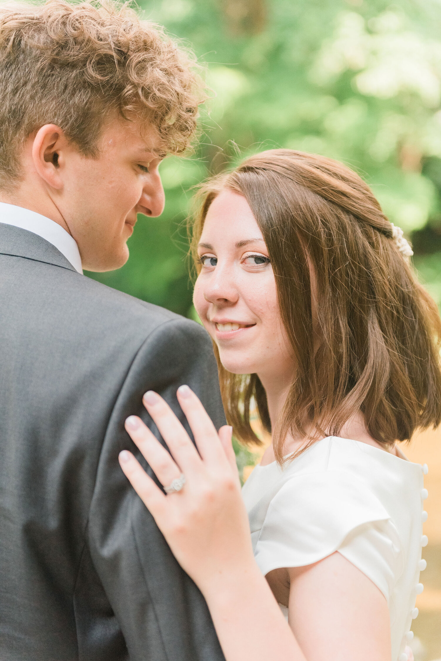  A bride peeks over her groom’s shoulder and shows her lovely wedding ring on their wedding day in Philadelphia, PA. bride and groom pose idea wedding day in a park outdoor wedding photography wedding hair ideas groom outfit ideas wedding ring ideas 
