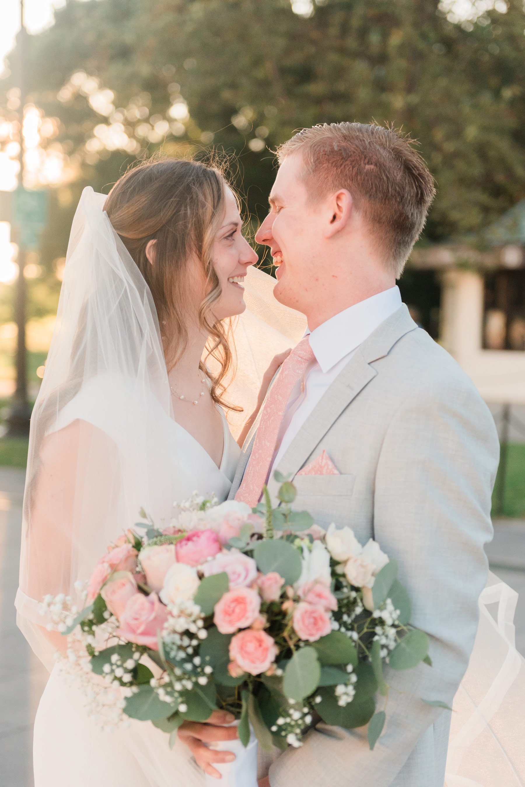    The Washington DC bride and groom smile at each other holder her pink rose bouquet. #washingtondcphotography #washingtondctemple #washingtondcwedding #dcweddingphotographer #washingtondctemplewedding #eastcoastweddingphotographer #sunsetbridals #m