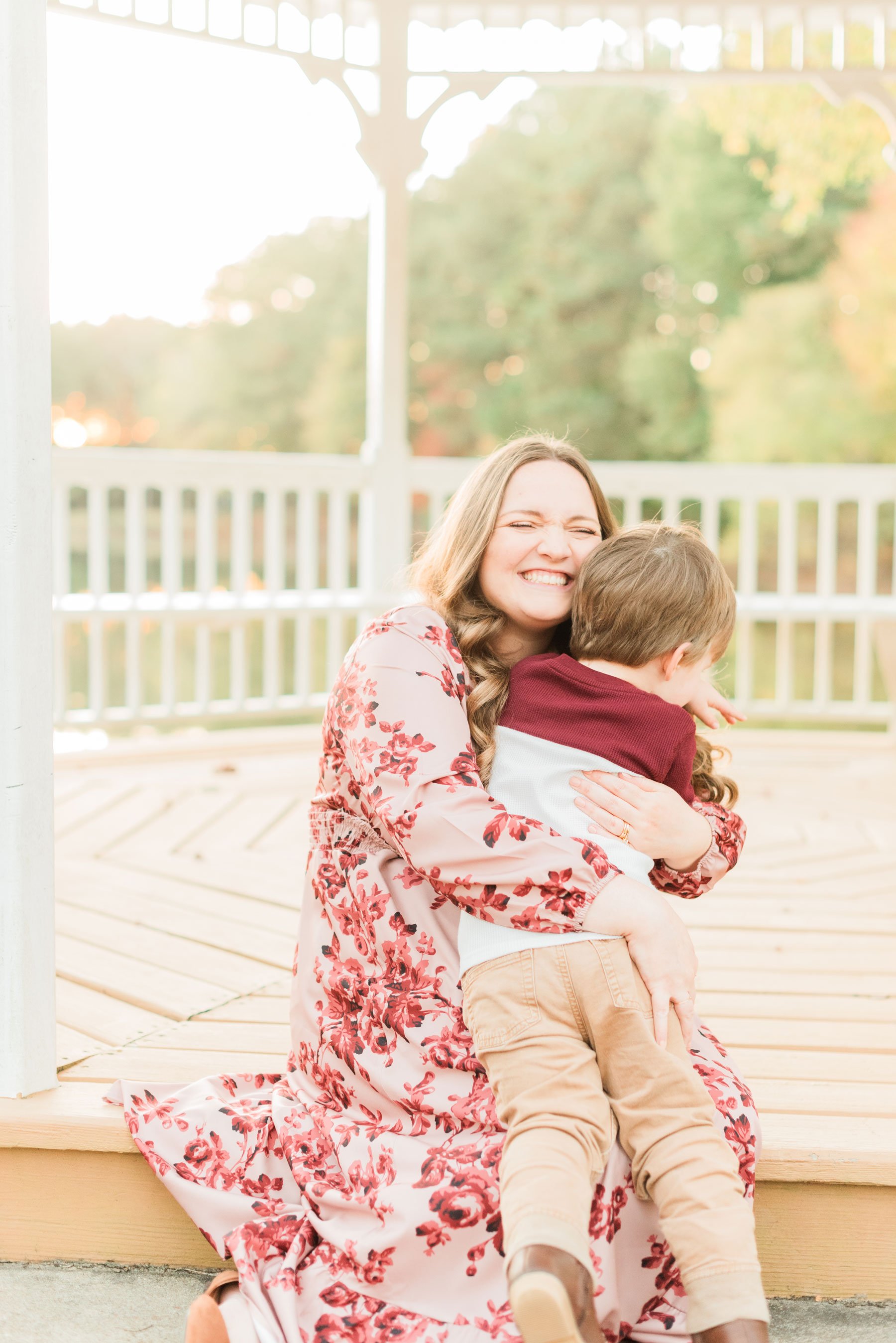  A mother wearing a floral pink and red dress gets a big hug from her two-year-old son in a gazebo in the fall. Georgia Maternity photos mother son hug&nbsp; #familymaterintyphotos #pregnacyphotoshoot #familyof3 #peachtreecity #atlantafamilyphotograp
