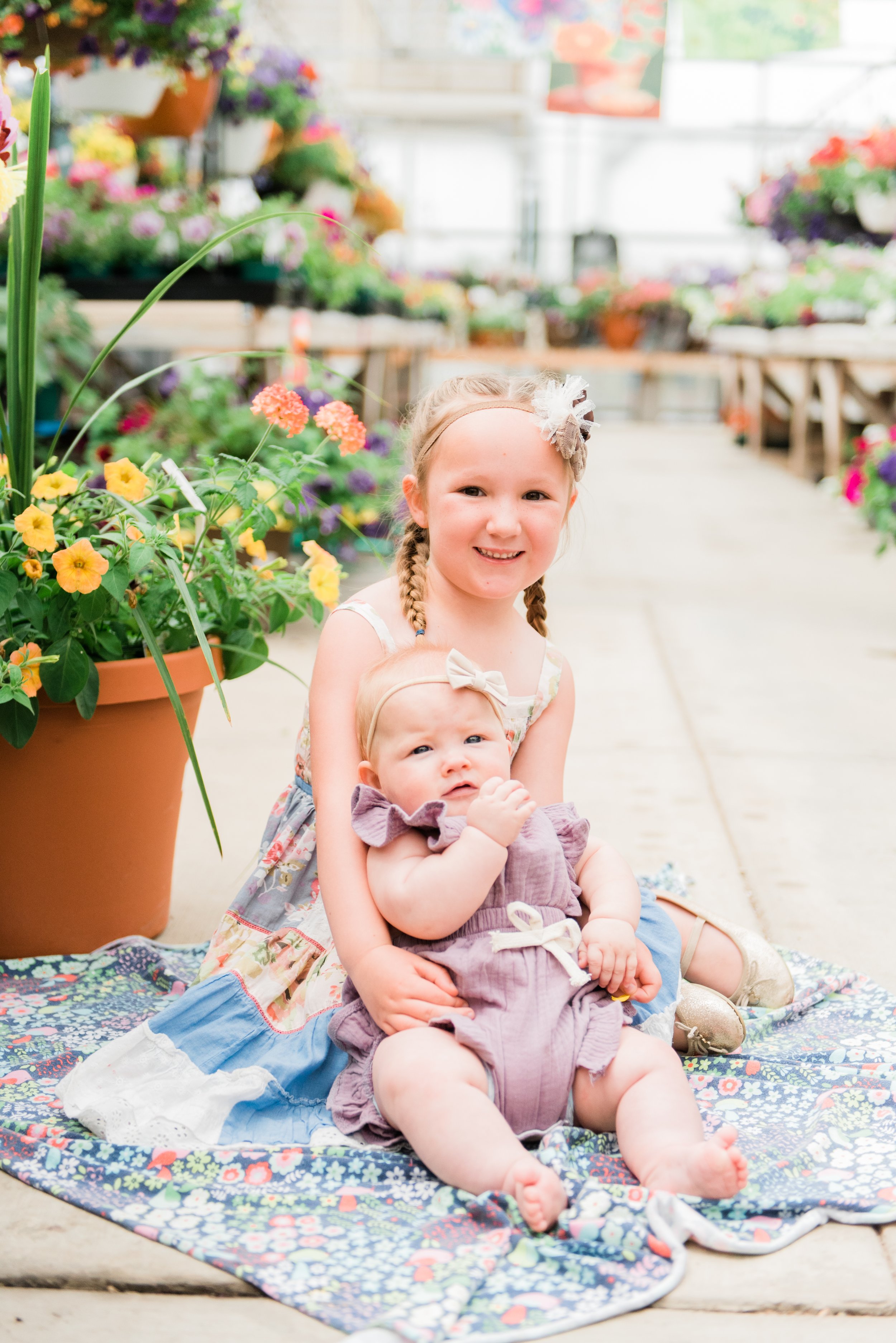  A big sister cradles her baby sister in a floral nursery full of colorful flowers. Sisters floral nursery lavender outfits Jacquie Erickson #babysister #gardennursery #bigsister #summerphotoshoot #jacquieerickson #georgiaphotographer 