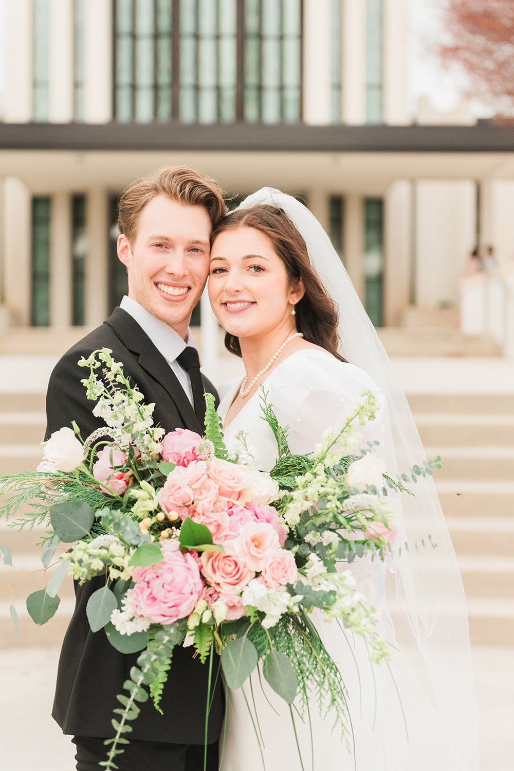  A bride holding a large bouquet of pink and white flowers, and her groom smile together in front of the Atlanta, Georgia LDS temple Professional wedding photographer #peachtreecity #weddingphotographer #ldsweddings #atlantatemple #jacquieerickson 