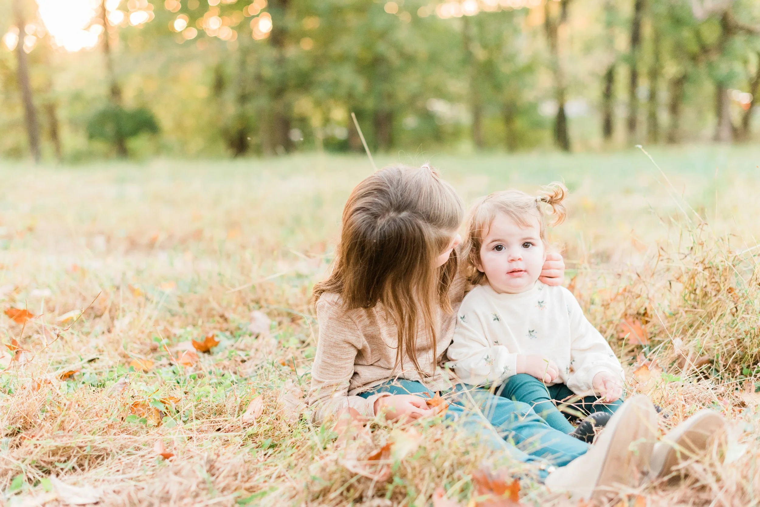 Two sisters dressed in pink sit together in a meadow. The older sister is looking at her baby sister. Photos of young children capturing memories mom tog course family photos  #jacquieerickson #takepicturesofyourkids #momtog #captureeverydaymoments