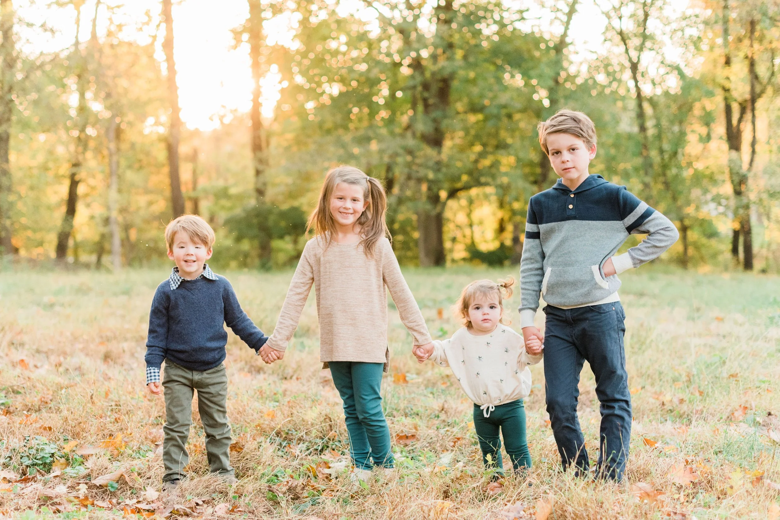 Four young siblings stand together holding hands in a forest meadow at dusk. Photos of young children capturing memories mom tog course #jacquieerickson #takepicturesofyourkids #momtog #captureeverydaymoments
