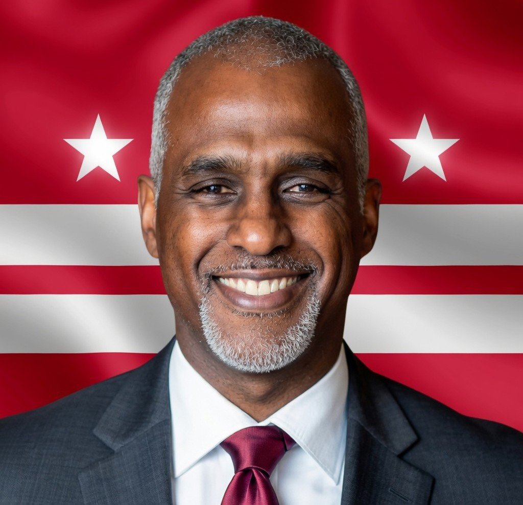 Talib Karim Muhammad, in a suit and tie, standing in front of a DC flag backdrop.