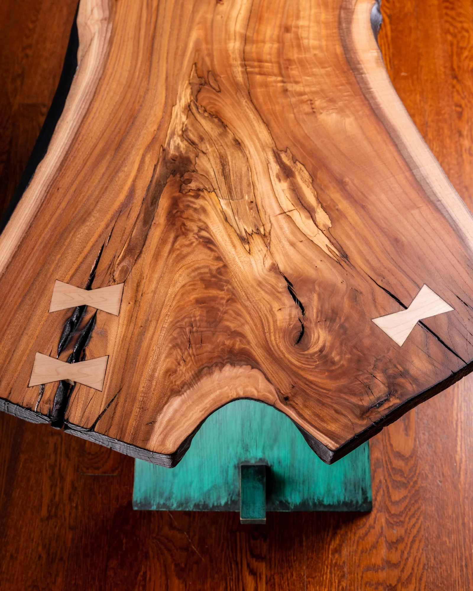 Close-up of a wooden table with a natural, live edge and a colorful finish, supported by a teal-colored base.