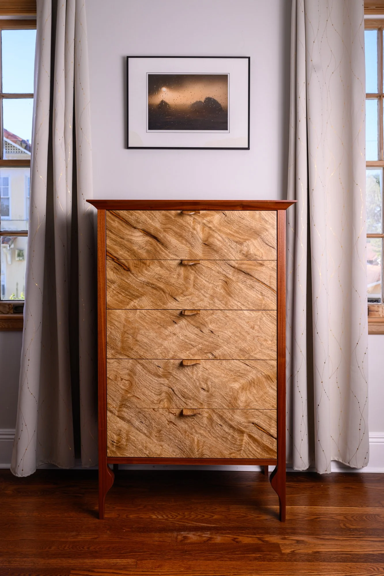 A wooden dresser with multiple drawers standing in a room, with two windows and curtains on either side and a framed picture hanging above it.