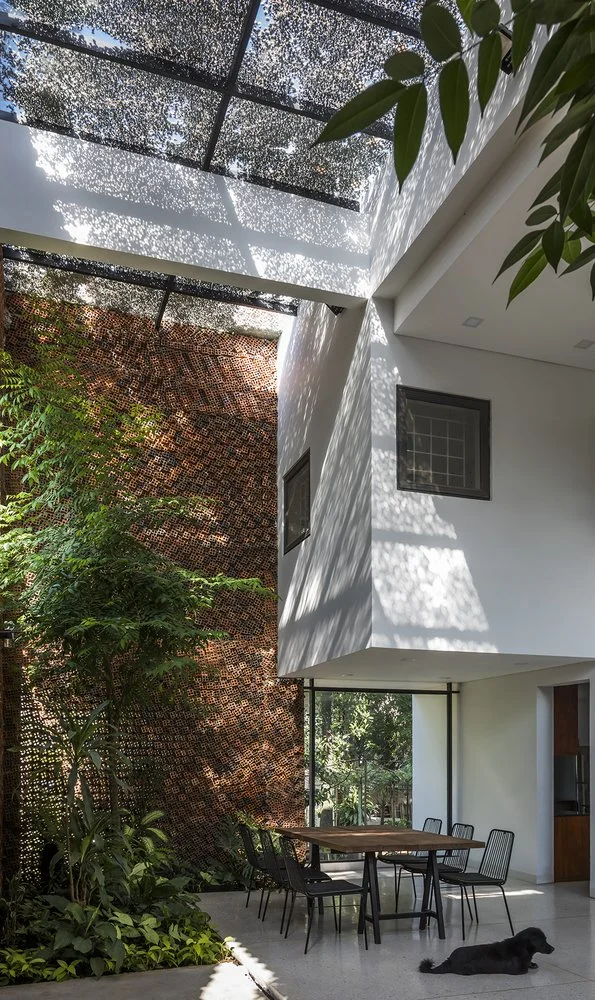 位於越南的Wall House,由CTA | Creative Architects設計,是一棟現代住宅的室內中庭,擁有一面高聳的磚牆、茂密的綠色植物和一棵樹。透明的屋頂允許陽光灑落,在白色牆壁和地板上形成光影圖案。下方擺放著一張餐桌和椅子,角落還趴著一隻黑狗在休息。