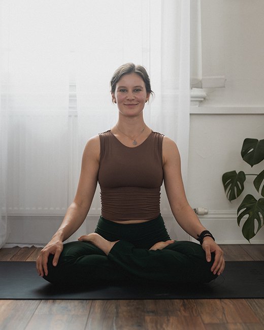 A young woman practicing yoga indoors, sitting cross-legged on a yoga mat, with a background of white curtains and a plant.