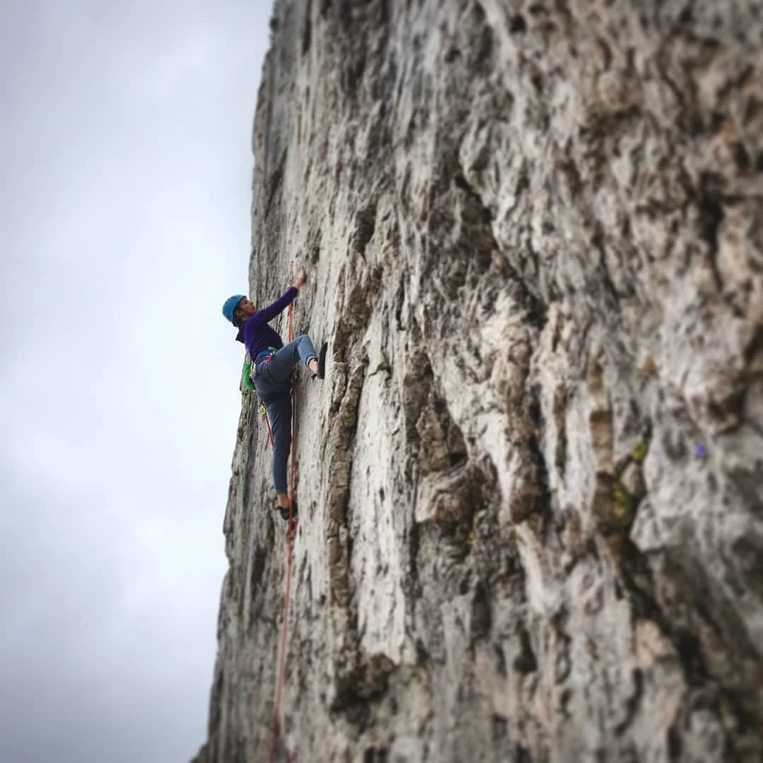 A white woman rock climbing on a steep, tall grey rocky crag. She is wearing a purple top, light grey trousers and is looking up to see where to go next.
