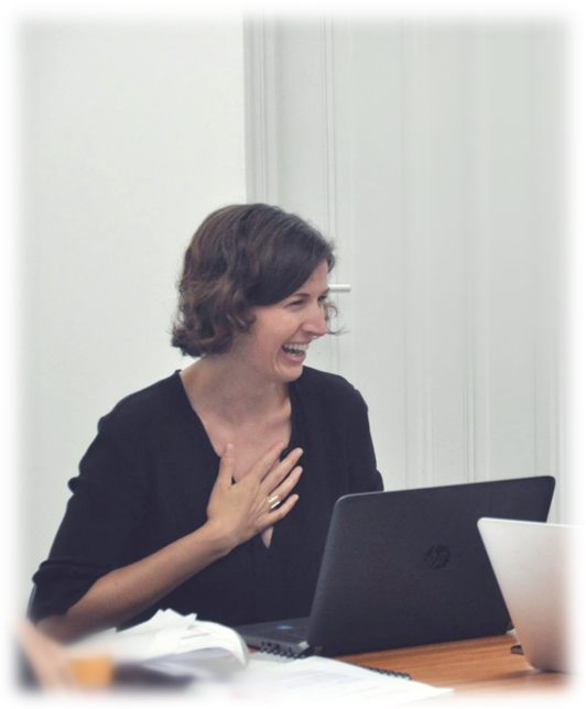 Woman with short brown hair, smiling and touching her chest, seated at a table with laptops and papers.