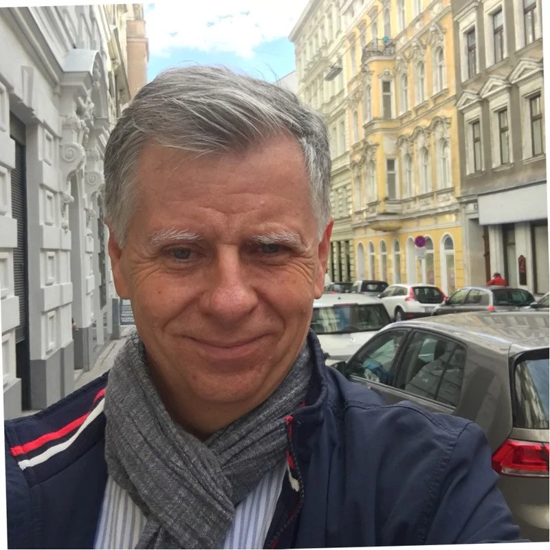 Michael Fembek, a man with gray hair and a scarf on a city street with colorful buildings and parked cars in the background.