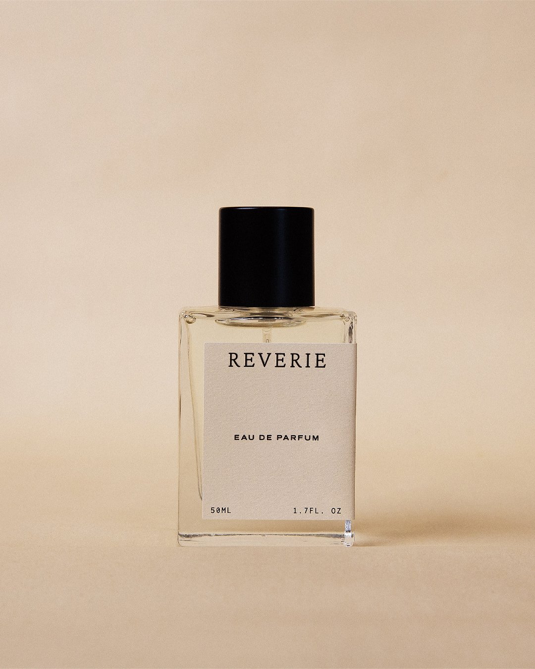 A clear glass perfume bottle with a black cap and a white label reading "REVERIE Eau de Parfum."