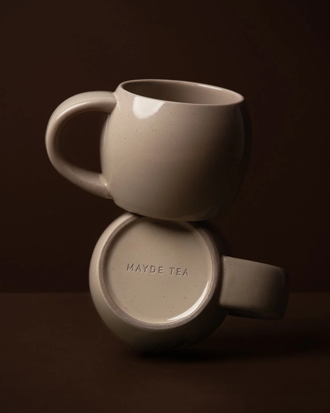 Two white ceramic mugs stacked with one on top of the other on a brown background. The bottom mug has the words 'MAYDE TEA' embossed on its base.