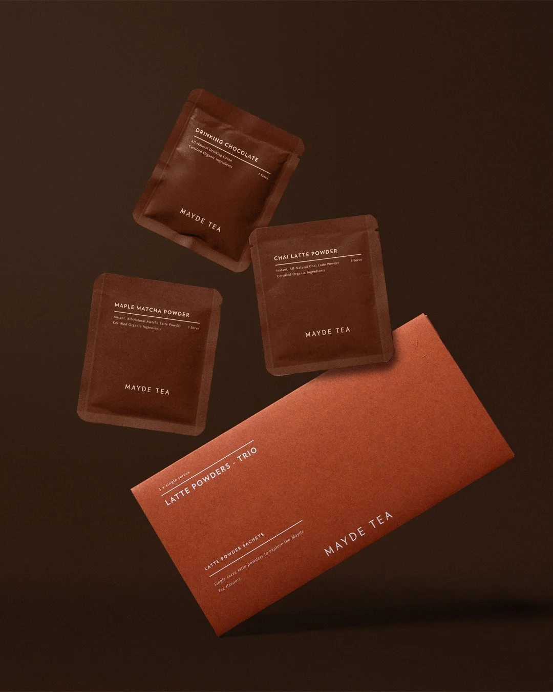Set of Mayde Tea latte powder sachets and a box, including Drinking Chocolate, Chai Latte Powder, and Maple Matcha Powder on a dark background.