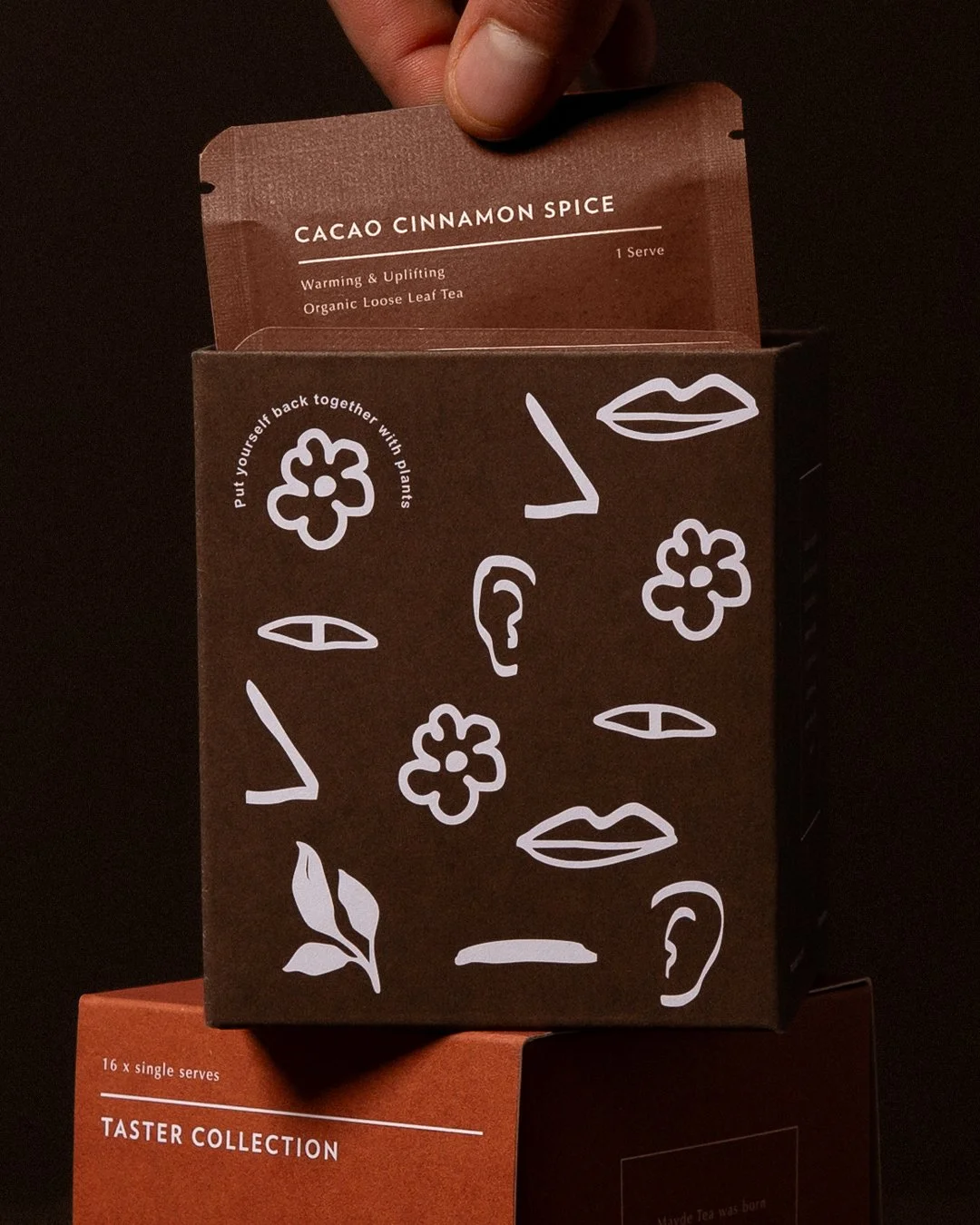 A person holding a brown packet of Cacao Cinnamon Spice tea, which is partially pulled out from a black box decorated with white line drawings of faces, lips, leaves, and flowers.